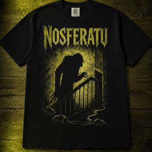 Può includere: Una t-shirt nera a maniche corte con una stampa grafica gialla della silhouette di Nosferatu che sale una scala con dei ratti alla base. Il testo NOSFERATU è stampato in un carattere giallo invecchiato sopra la figura centrale.