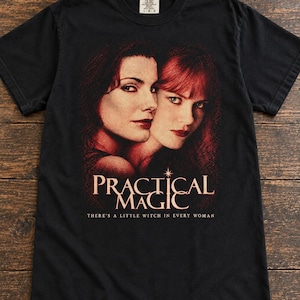 May include: A black short-sleeved t-shirt featuring a graphic print of two individuals with red-toned hair and skin. Below the faces, the text reads "PRACTICAL MAGIC" and "THERE'S A LITTLE WITCH IN EVERY WOMAN" in a light-coloured serif font.