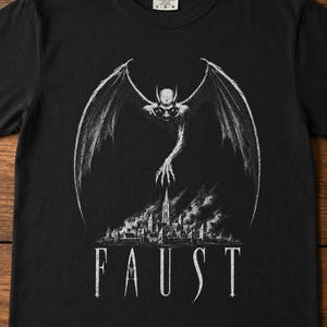 Puede incluir: Una camiseta negra de manga corta con un estampado gráfico blanco de una criatura alada sobrevolando una ciudad con humo. La palabra FAUST está impresa en letras mayúsculas blancas en la parte inferior. Etiqueta de cuello Comfort Colors.