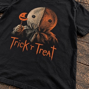 Pode incluir: Uma t-shirt preta de manga curta com um gráfico de uma personagem de saco de serapilheira com olhos de botão a segurar um chupa-chupa laranja. Abaixo da personagem, o texto Trick 'r Treat está impresso em laranja. Etiqueta Comfort Colors.