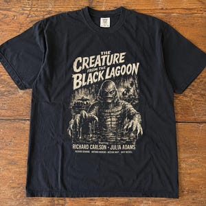 May include: A black short-sleeve t-shirt featuring a graphic print of The Creature from the Black Lagoon. The design includes the movie title, an illustration of the creature emerging from water, and the names of actors Richard Carlson and Julia Adams.