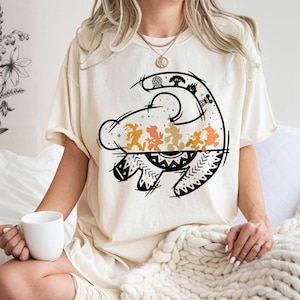 May include: A cream-colored short-sleeve t-shirt featuring a black line-art graphic of a stylized lion silhouette. Inside the lion shape are orange and yellow silhouettes of cartoon characters and park landmarks, accented with small stars.