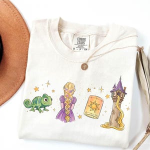 Peut inclure: Un t-shirt à manches courtes de couleur crème avec une impression graphique représentant un caméléon vert, un personnage aux longs cheveux blonds tressés portant une robe violette, une lanterne flottante et une haute tour en pierre.