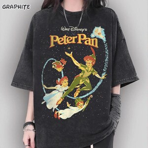 Puede incluir: Una camiseta gris carbón con efecto lavado al ácido que presenta un estampado gráfico de Peter Pan, Wendy, John y Michael Darling volando. El texto dice Walt Disney's Peter Pan. El diseño incluye un rastro de polvo de hadas sobre un fondo estrellado.