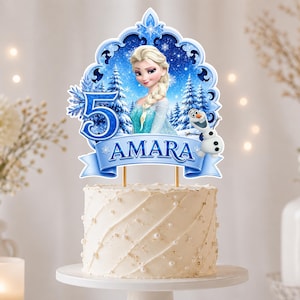 May include: A cake topper featuring a blue ornate frame with a blonde character in a blue dress, a snowman, a large blue number 5, and the name AMARA in blue text. The topper is mounted on two thin sticks and sits atop a white frosted cake.