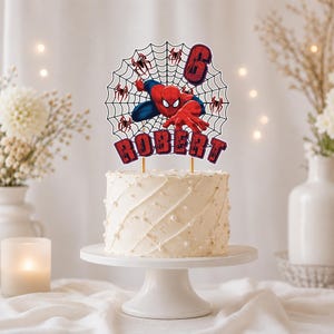 May include: A cake topper featuring a red and blue superhero character in a black spiderweb design. The topper includes the number 6 and the name ROBERT in red block letters with black outlines. It is mounted on two thin sticks atop a white cake.