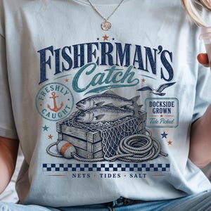 May include: A light blue t-shirt featuring a graphic print with the text "FISHERMAN'S Catch", "FRESHLY CAUGHT", "DOCKSIDE GROWN", "Tide Picked", and "NETS TIDES SALT". The design includes two fish in a wooden crate, a fishing net, and an anchor.