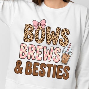 Könnte beinhalten: Ein weißes Langarm-Sweatshirt mit einem Grafikdruck und dem Text "BOWS BREWS & BESTIES". Das Wort "BOWS" ist in Leopardenmuster mit einer rosa Schleife gehalten, "BREWS" in einem rosa-weißen Schachbrettmuster und "BESTIES" in Braun.