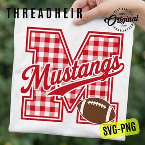 May include: A white t-shirt featuring a large letter M with a red and white gingham pattern. The word Mustangs is written in red script across the letter. A brown football with white laces is positioned at the bottom right of the letter M.