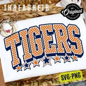 May include: A white t-shirt featuring the word TIGERS in large, orange block letters with white polka dots and a dark blue outline. Below the text is a row of alternating orange and dark blue stars. The text THREADHEIR appears at the top.