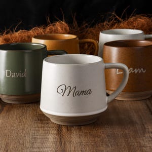 Puede incluir: Una colección de tazas de cerámica en varios colores, incluyendo verde oliva, amarillo mostaza, blanco y marrón. Cada taza presenta un acabado liso con una textura moteada y una base de arcilla natural. La taza blanca está grabada con la palabra Mama.