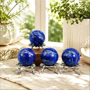 Pode incluir: Quatro esferas de lápis-lazúli polido, cada uma com uma coloração azul profunda e inclusões de pirita dourada. Cada esfera é exibida num suporte de tripé decorativo de metal prateado com pernas curvas e ornamentadas.