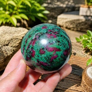 May include: A polished spherical stone featuring a vibrant mix of deep magenta-red ruby inclusions embedded within a dark green zoisite matrix, displaying natural black mineral speckles throughout the smooth, rounded surface of the orb.