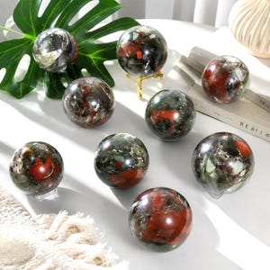 May include: A collection of eight polished stone spheres featuring a mottled mix of dark green, black, and grey minerals with prominent, smooth, rounded inclusions of deep red jasper throughout the surface of each individual orb.