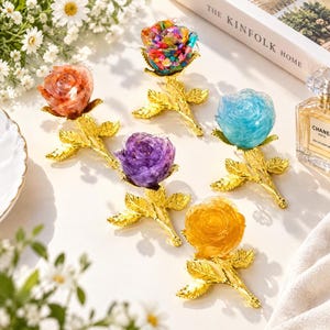 Könnte beinhalten: Fünf dekorative Rosenornamente, jedes mit einem durchscheinenden, farbigen Harzblütenkopf an einem metallisch goldenen Stiel mit Blättern. Die Rosen sind orange, mehrfarbig, lila, hellblau und gelb und auf einer Oberfläche angeordnet.