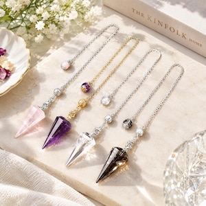 May include: Four crystal pendulums with pointed gemstone tips in rose quartz, amethyst, clear quartz, and smoky quartz. Each pendulum hangs from a delicate metal chain featuring decorative beads and a small charm at the end of the chain.