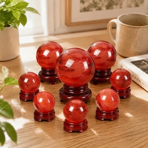 May include: A collection of nine smooth, translucent red spheres with swirling internal patterns, each resting on a dark brown circular wooden stand. The spheres vary in size, with one large central piece surrounded by eight smaller ones.