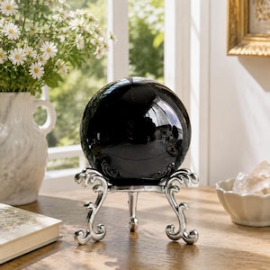 Può includere: Una sfera nera opaca e lucida che poggia su un supporto decorativo in metallo color argento con tre gambe curve e ornate. La superficie liscia e riflettente della sfera cattura la luce e mostra un riflesso distorto dell'ambiente.