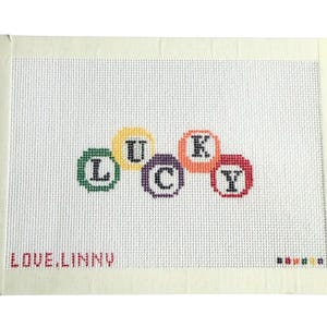 May include: A white needlepoint canvas featuring the word LUCKY stitched in black letters inside colorful circular borders of green, yellow, purple, orange, and red. The text LOVE,LINNY is stitched in red in the bottom left corner.