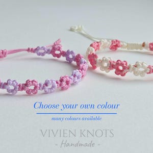 May include: Two macrame bracelets featuring a repeating flower pattern. One bracelet is made of pink and light purple cord, while the other is white and pink. Text reads: Choose your own colour, many colours available, VIVIEN KNOTS Handmade.