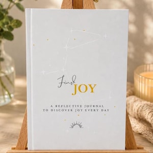 May include: A white journal titled Find JOY A REFLECTIVE JOURNAL TO DISCOVER JOY EVERY DAY. The cover features a light grey constellation pattern with small gold stars and a sun icon at the bottom, displayed on a small wooden easel stand.