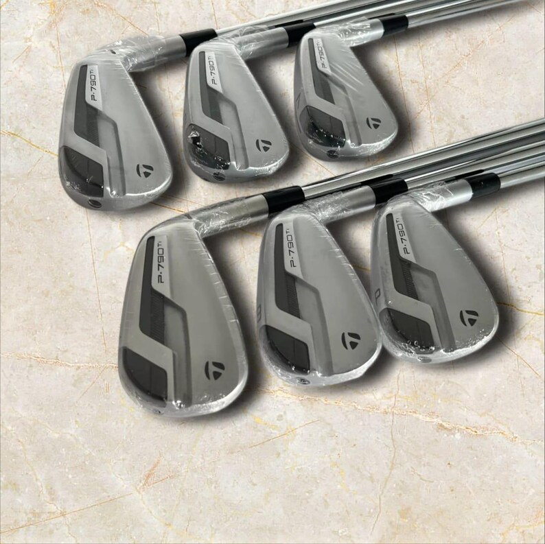 Puede incluir: Un juego de seis hierros de golf TaylorMade P-790 Ti con cabezas de color gris met&aacute;lico y detalles en negro. Cada cabeza de palo est&aacute; parcialmente envuelta en pl&aacute;stico protector transparente, mostrando el nombre P-790 Ti y el logo de TaylorMade.