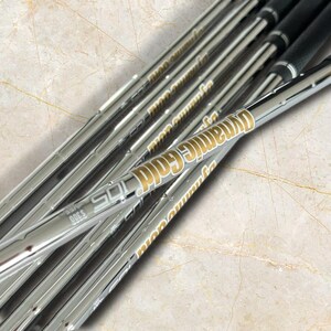 Puede incluir: Un conjunto de varas de palo de golf de acero con acabado cromado que presentan texto de color dorado que dice Dynamic Gold 105 y S300. Las varas est&aacute;n dispuestas en diagonal, mostrando una superficie met&aacute;lica reflectante con marca clara.