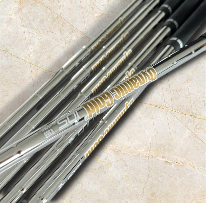 Puede incluir: Un conjunto de varas de palo de golf de acero con acabado cromado que presentan texto de color dorado que dice Dynamic Gold 105 y S300. Las varas est&aacute;n dispuestas en diagonal, mostrando una superficie met&aacute;lica reflectante con marca clara.
