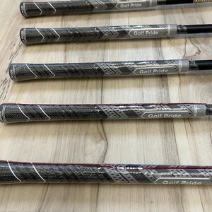 May include: A collection of golf club grips featuring a textured black and grey pattern with white chevron accents. Each grip is wrapped in clear protective plastic and displays the Golf Pride logo in white text on a grey rectangular background.