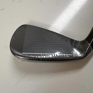 May include: A black golf iron club head featuring a series of horizontal grooves on the face. The club head is encased in clear protective plastic film, showing a smooth, dark finish and a distinct white line along the bottom edge of the face.