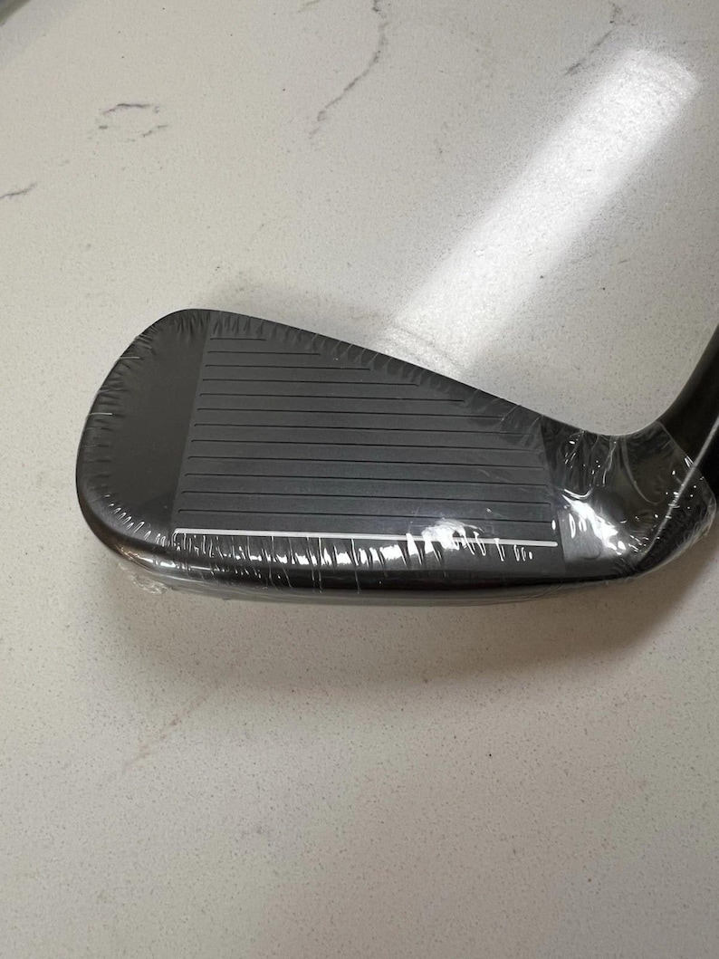 May include: A black golf iron club head featuring a series of horizontal grooves on the face. The club head is encased in clear protective plastic film, showing a smooth, dark finish and a distinct white line along the bottom edge of the face.