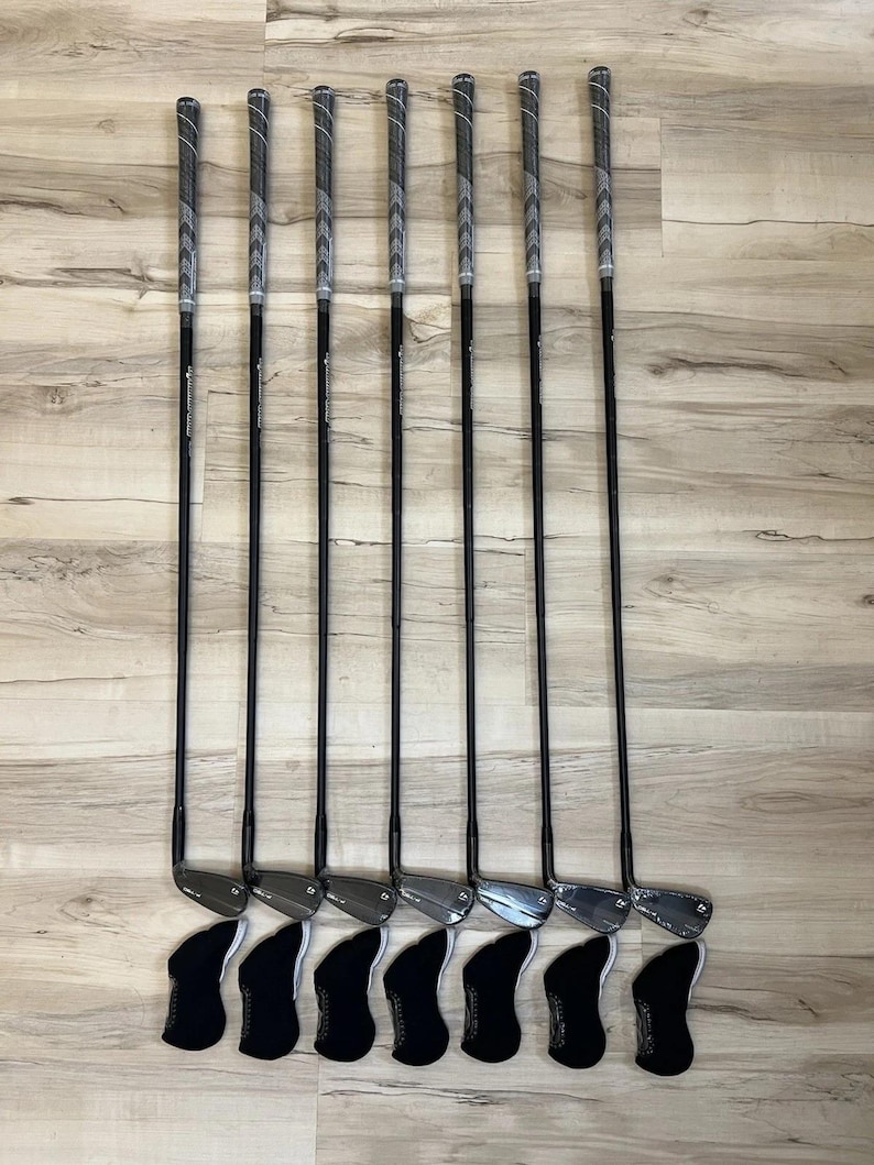 May include: A set of seven golf irons with black shafts and textured grey grips, each featuring a metallic club head and a black protective headcover positioned at the base. The clubs are arranged in a vertical row, showcasing a consistent design.