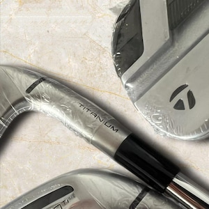 Puede incluir: Un juego de hierros de golf de tono plateado envueltos en pl&aacute;stico protector transparente. Las cabezas de los palos presentan la palabra TITANIUM grabada en el cuello y un logotipo negro de TaylorMade en la parte posterior con acabado met&aacute;lico.