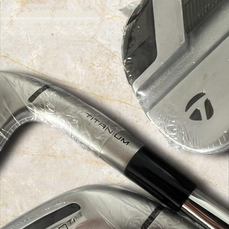 Puede incluir: Un juego de hierros de golf de tono plateado envueltos en pl&aacute;stico protector transparente. Las cabezas de los palos presentan la palabra TITANIUM grabada en el cuello y un logotipo negro de TaylorMade en la parte posterior con acabado met&aacute;lico.