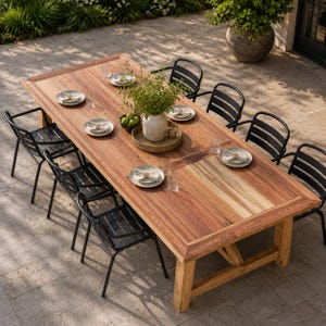 May include: A rectangular wooden dining table with a natural grain finish, surrounded by six black metal chairs with slatted backs. The table is set with six white plates, napkins, and a central round tray holding a green plant in a ceramic vase.
