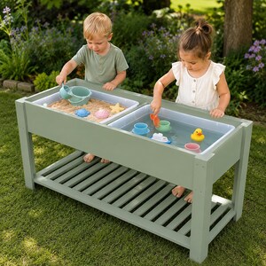 Peut inclure: Une table sensorielle en bois vert sauge dotée de deux bacs en plastique amovibles, l'un rempli de sable et l'autre d'eau. La table comprend une étagère inférieure à lattes pour le rangement et divers jouets en plastique colorés, dont un canard jaune.