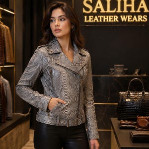 Könnte beinhalten: Eine graue und schwarze Leder-Motorradjacke mit Schlangenledermuster, Reverskragen, asymmetrischem Reißverschluss und einem Gürtel mit Schnalle in der Taille. Die Jacke hat lange Ärmel und Reißverschlusstaschen und wird vor dem Schild SALIHA LEATHER WEARS präsentiert.