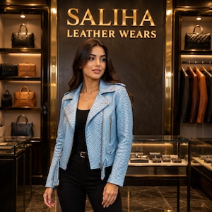 May include: A light blue motorcycle jacket with a snakeskin texture, featuring a notched collar, silver-tone metal zippers, and a belted waist. The jacket is worn over a black top and black pants, set against a store background with the text SALIHA LEATHER WEARS.