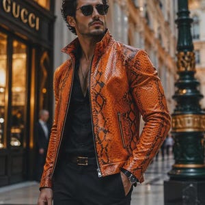 Peut inclure: Une veste orange vif présentant un motif texturé en peau de serpent avec des accents noirs, un col montant et une fermeture éclair frontale. La veste est portée sur une chemise noire boutonnée et un pantalon noir, avec une ceinture et une montre.