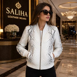 May include: A white bomber jacket featuring a black and white snakeskin pattern, a silver front zip, and solid white ribbed cuffs and collar. The jacket is worn over a plain white top, paired with black trousers and dark-tinted sunglasses.