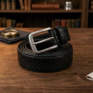 May include: A black belt with a textured, snakeskin-like pattern and a brushed silver-tone metal buckle. The belt is coiled into a circular shape, showcasing the detailed scale-like surface texture and the sturdy, rectangular metal pin buckle.