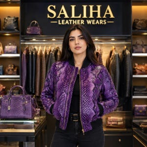 May include: A vibrant purple bomber jacket featuring a textured snakeskin pattern, a black ribbed collar, cuffs, and hem, and a central gold-tone zip. The jacket is worn over a black top and dark jeans in a retail store with the text SALIHA LEATHER WEARS.