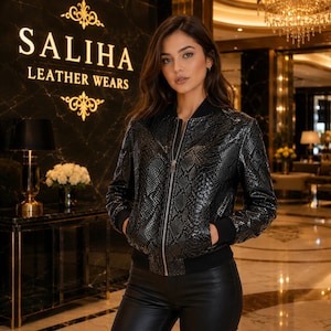 May include: A black bomber jacket with a snakeskin texture, featuring a front silver zip closure, black ribbed cuffs, and a black ribbed hem. The jacket is worn over black trousers. The background displays the text SALIHA LEATHER WEARS.