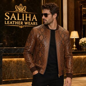 Puede incluir: Una chaqueta bomber de cuero marrón con estampado de piel de serpiente, cuello, puños y dobladillo de canalé marrón oscuro. La chaqueta tiene cierre de cremallera frontal y se lleva sobre una camiseta negra. El texto SALIHA LEATHER WEARS es visible.