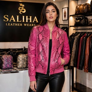 May include: A bright pink bomber jacket featuring a textured snakeskin pattern, a front zip closure, and solid pink ribbed cuffs and collar. The jacket is worn over a black top and black trousers, with a snake logo and text SALIHA LEATHER WEARS.