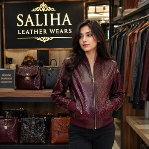 Peut inclure: Un blouson bomber bordeaux profond avec une texture imitation peau de serpent, doté d'une fermeture éclair frontale, de poignets côtelés et d'un ourlet côtelé. La veste est présentée dans un magasin avec une enseigne SALIHA LEATHER WEARS.
