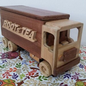 Può includere: Un camion giocattolo in legno con un vano di carico marrone scuro che riporta la scritta ROOT & TEA in lettere in rilievo. Il camion presenta una cabina in legno color beige chiaro, un tetto rettangolare, sei ruote piccole, una griglia frontale e finestrini.