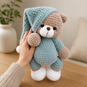 May include: A crocheted teddy bear toy made of soft, textured yarn. The bear has a light brown head and limbs, a white muzzle, and is wearing a teal blue sleeper suit with a matching long, pointed nightcap that features a white pom-pom at the end.
