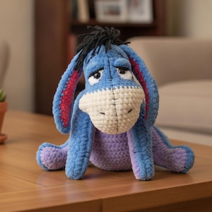 May include: A crocheted plush donkey toy featuring a blue body, a light cream-colored muzzle, and long ears with pink inner sections. The character has black yarn hair on top of its head, drooping eyes, and a soft, textured yarn construction.