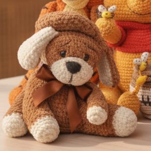May include: A brown and cream crocheted dog plush toy featuring a dark brown satin ribbon tied around its neck, floppy ears, and a dark nose. The toy is crafted with a textured yarn finish and is positioned in front of other crocheted figures.